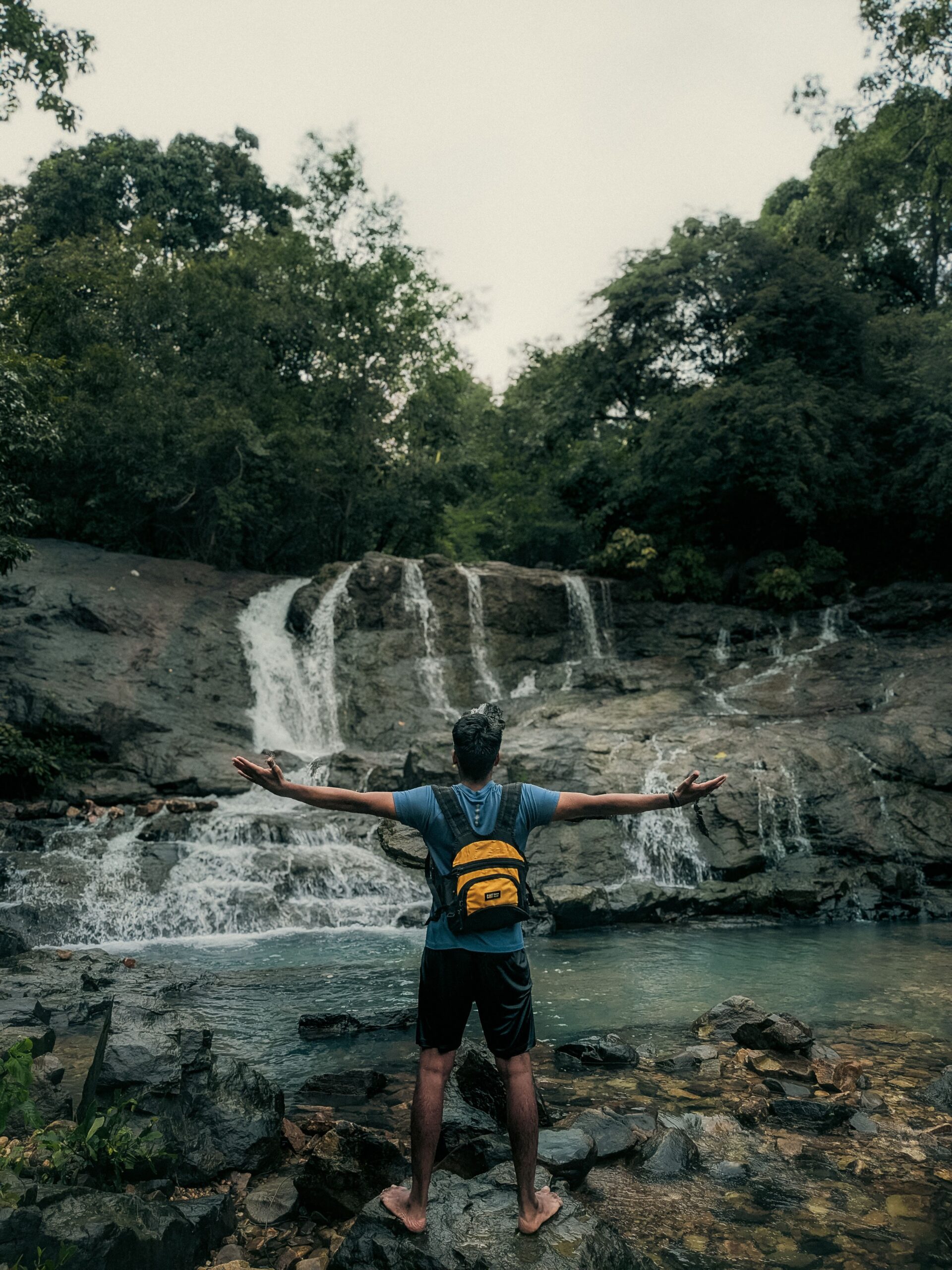 About Person with open arms stands at a serene waterfall surrounded by lush greenery.