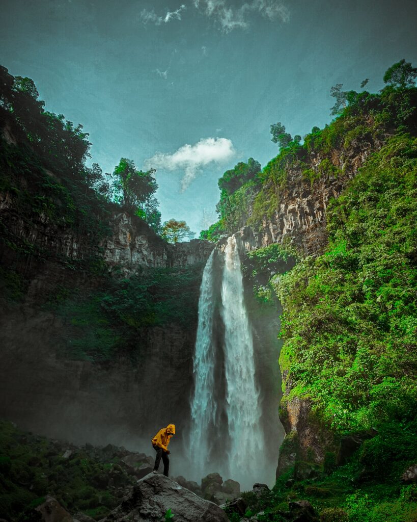 A man in a yellow hoodie explores a stunning waterfall surrounded by lush greenery, capturing nature's pristine beauty.